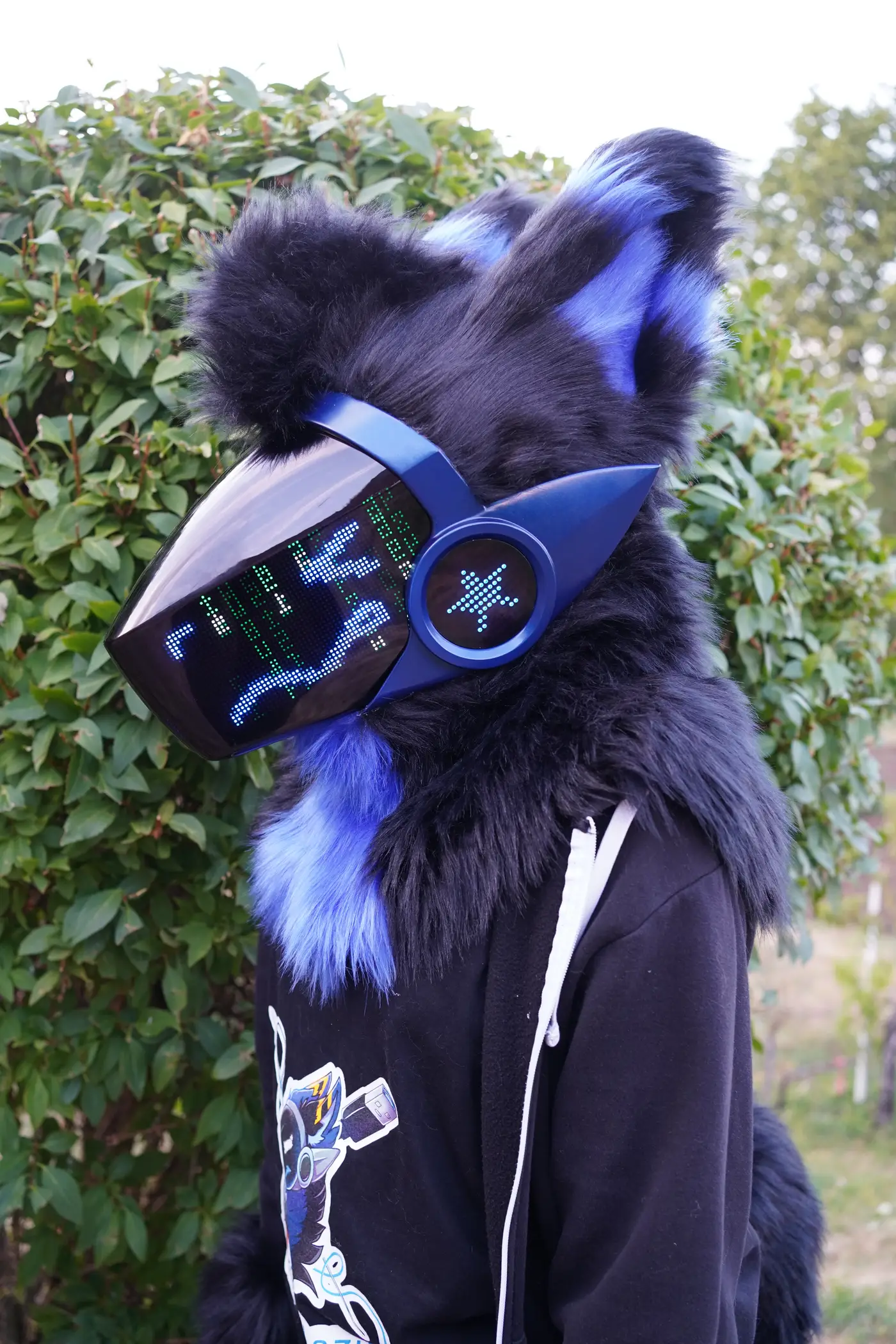 Close up of a black and blue Protogen head in front of a green hedge, with matrix style code on the LED visor