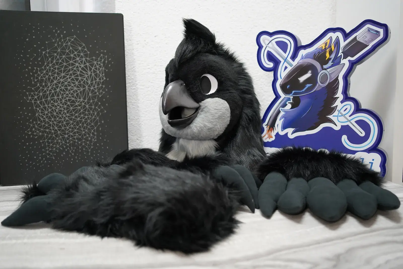 Black bird fursuit head with a gray beak and white eye patches, black hand paws and the Arget and Lazuli logo