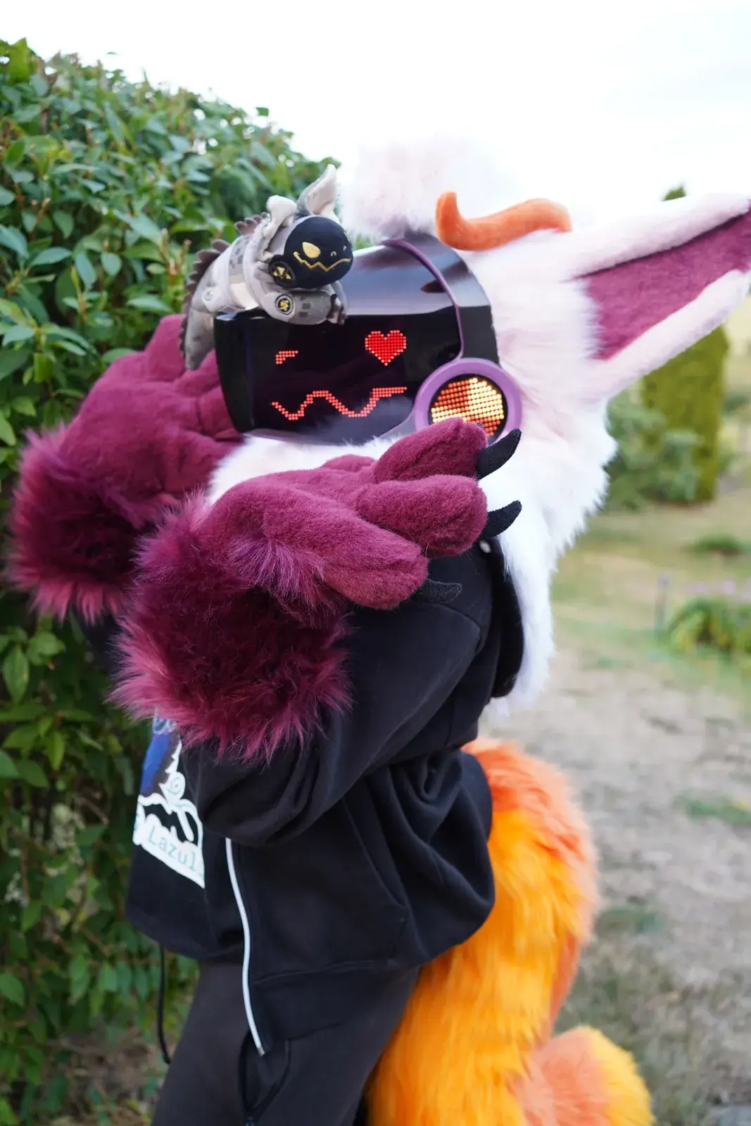 White and pink Protogen mini partial with orange horns, burgundy hand paws and an orange tail in the garden, a red heart on the LED visor