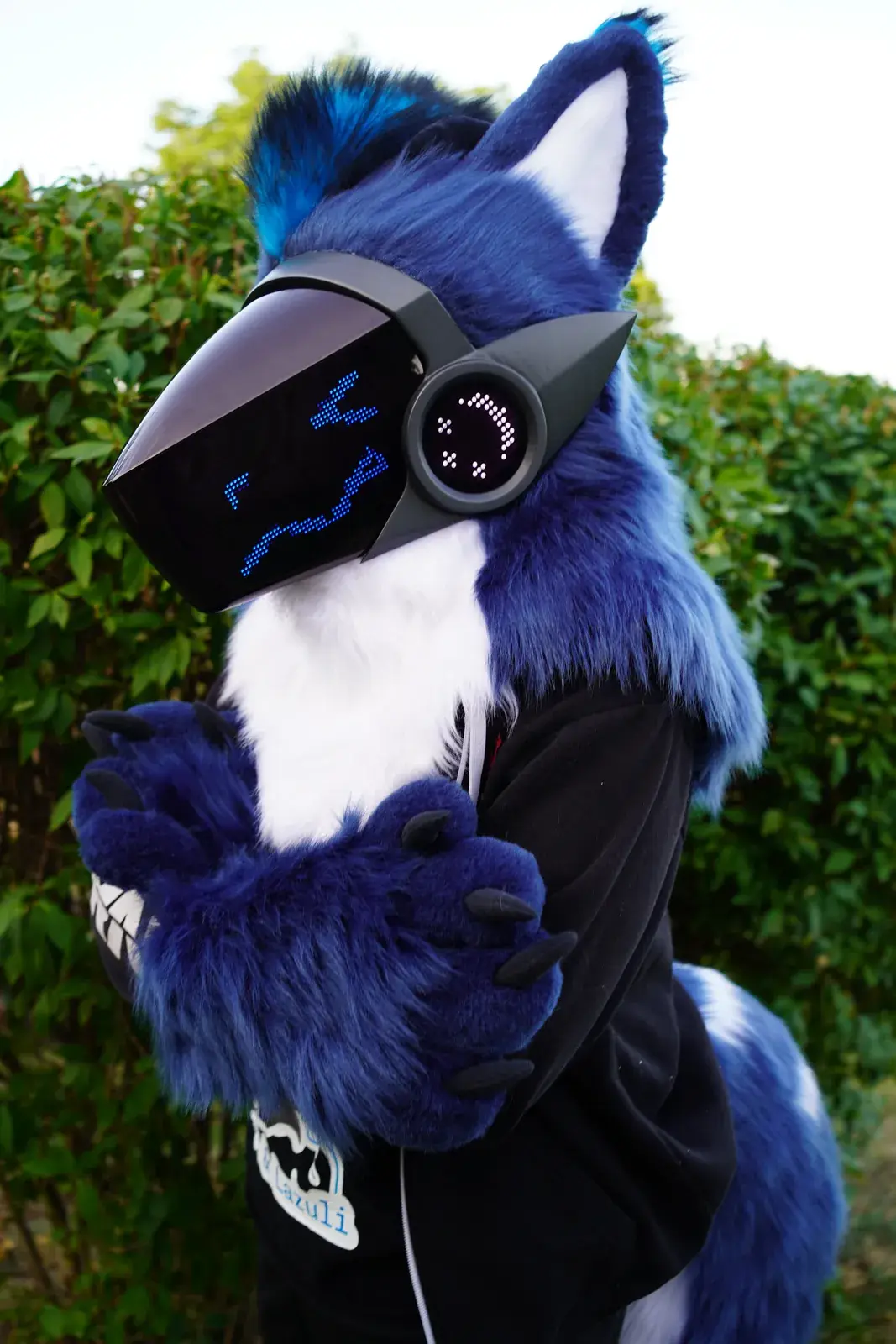 Blue and white Protogen mini partial by a garden hedge, blue hand paws with black claws and a blue smile on the LED visor
