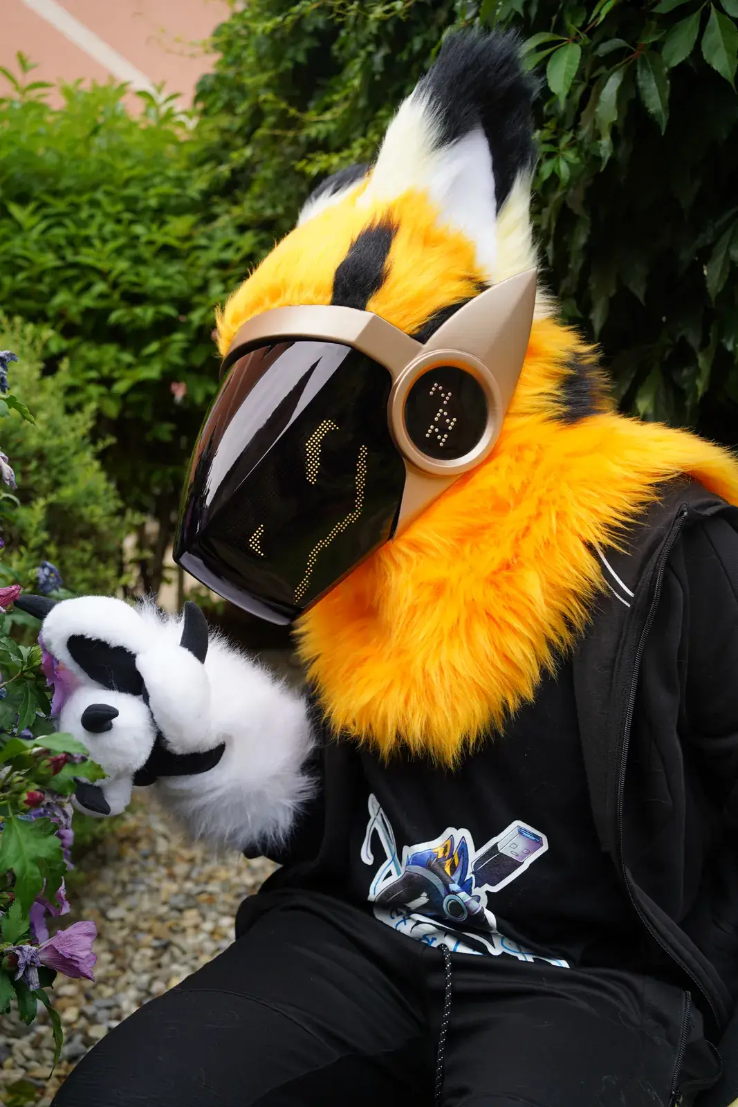 Orange Protogen mini partial sitting by purple flowers on a garden path, white hand paws with black pads