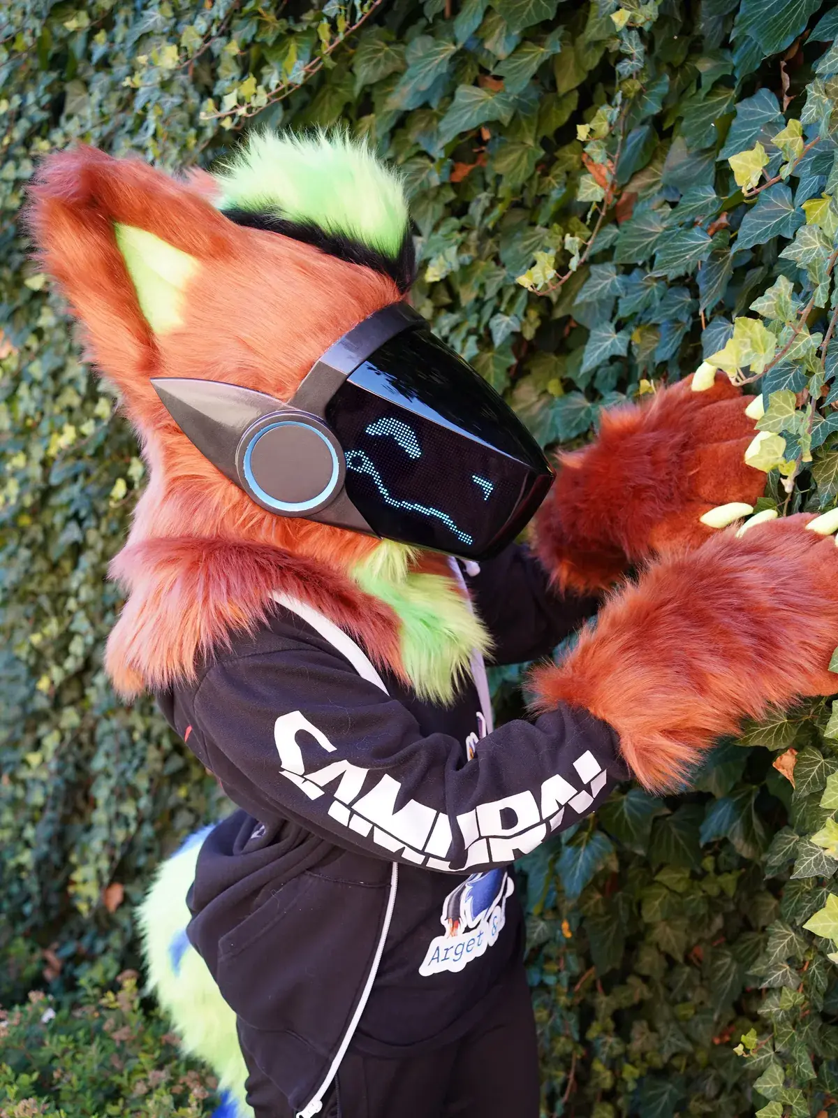 Rust brown Protogen partial with a lime green mane by an ivy wall, blue pixel face on the LED visor