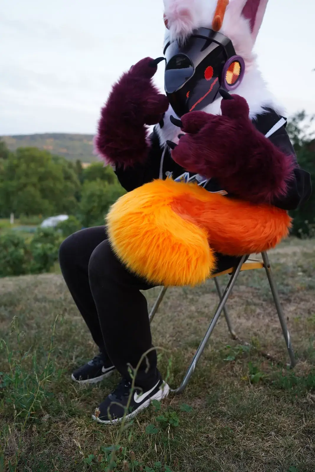 White and pink Protogen mini partial sitting on a folding chair in a meadow at dusk with its orange tail