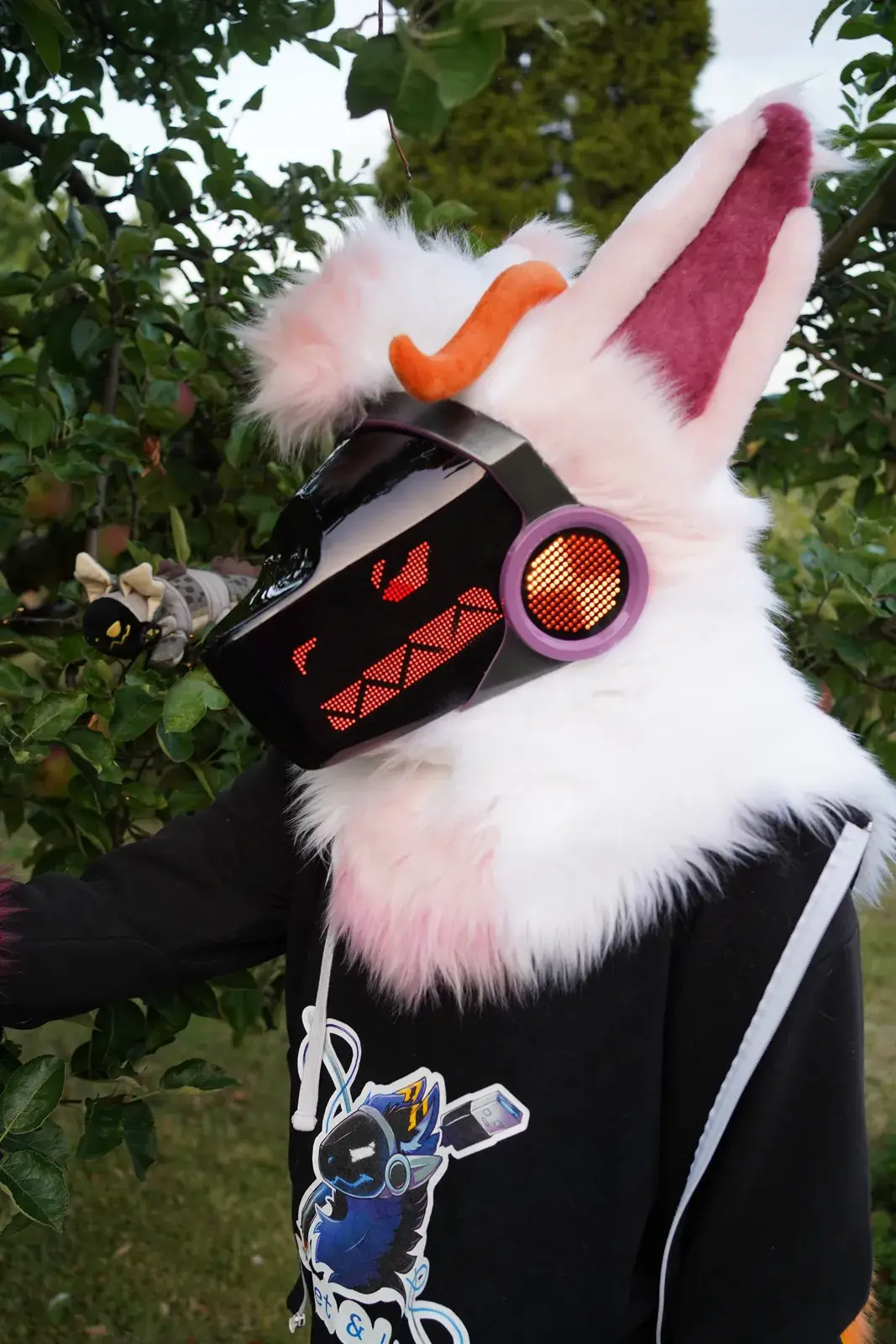 White and pink Protogen mini partial under an apple tree with a small figurine, narrowed red eyes on the LED visor