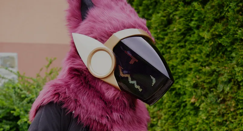 Burgundy Protogen mini partial with an orange face on the cream framed LED visor in front of a garden hedge, by Arget & Lazuli